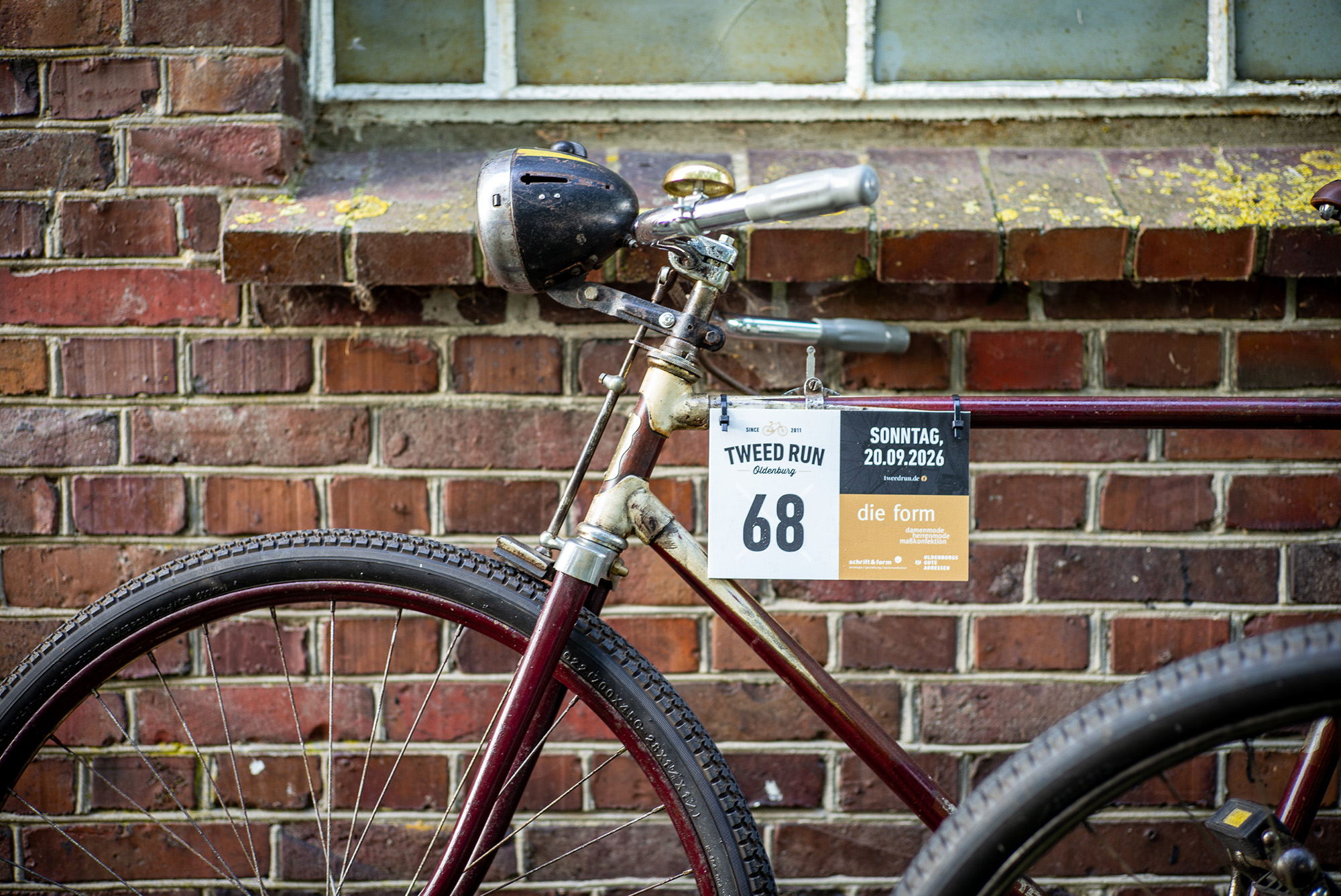 Altmodisches dunkelrotes Fahrrad mit dem Teilnehmerschild des Tweed Runs 2026 steht angelehnt an einer alten roten Backsteinwand.