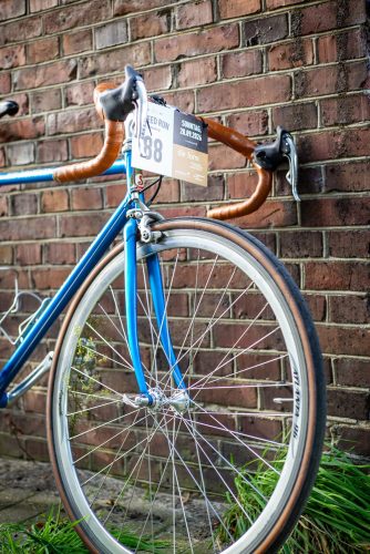 Hellblaues Vintage-Rennrad mit braunem Lenkerband und Starternummer 38 an der Backsteinmauer